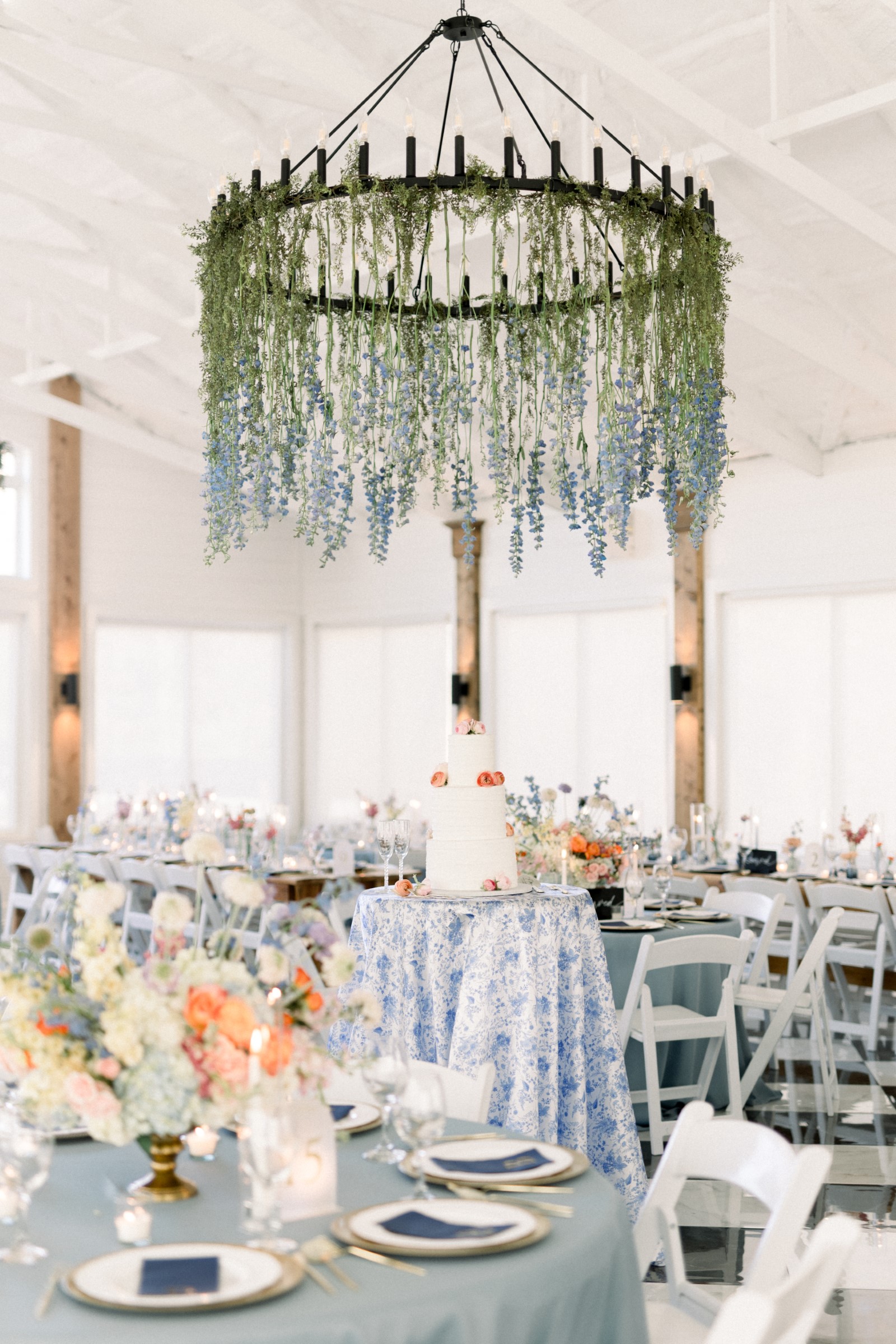 Wedding reception with a floral chandelier above a white cake and pastel-decorated blue tables