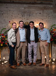 Close-up of a man with a woman in front of a brick wall; she wears a long floral dress.
