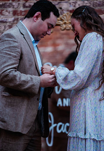 Four men in semi-formal attire standing arm-in-arm in front of a brick wall, flanked by flower arrangements and candles on the floor.
