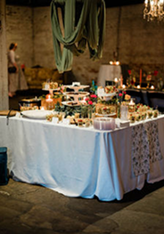 Dessert table in a rustic venue draped with fabric and a chandelier, displaying cakes, cupcakes, and floral accents