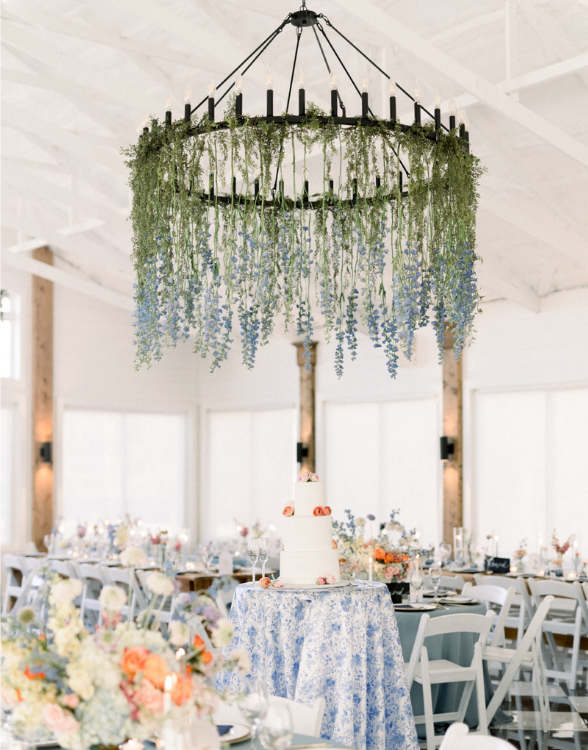 Wedding reception with a floral chandelier above a white cake and pastel-decorated blue tables